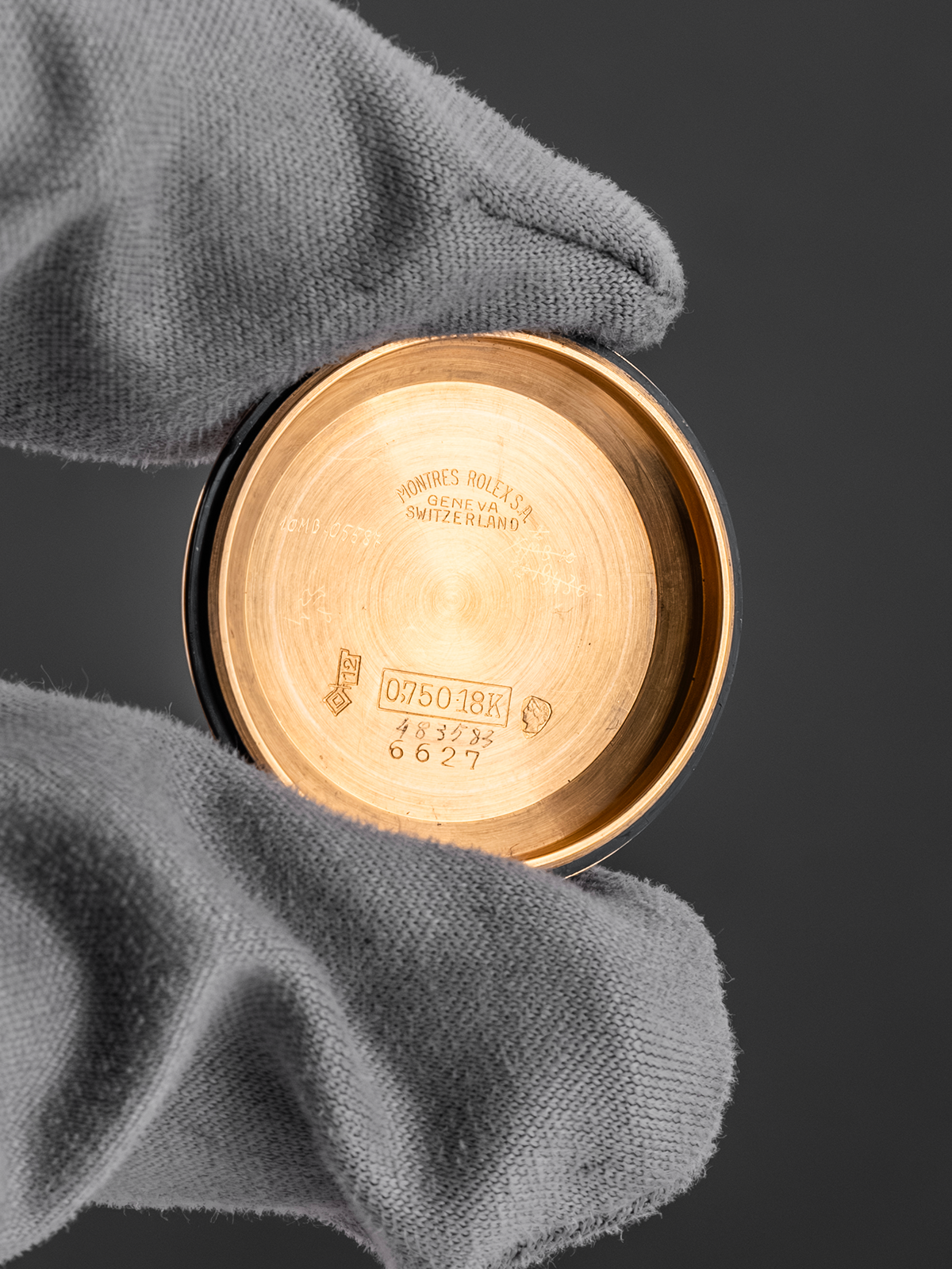 Gold watch case with brand engraving held by a gloved hand against a dark background
