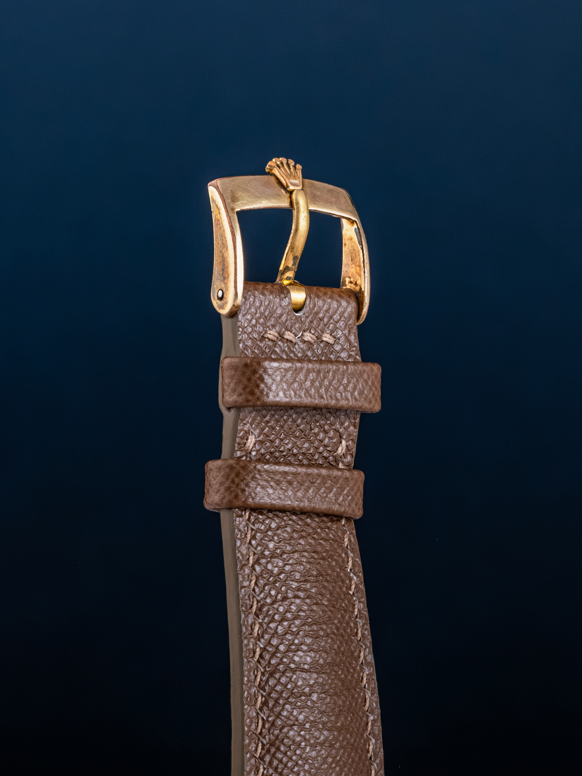 Brown leather watch strap with gold buckle on a dark blue background
