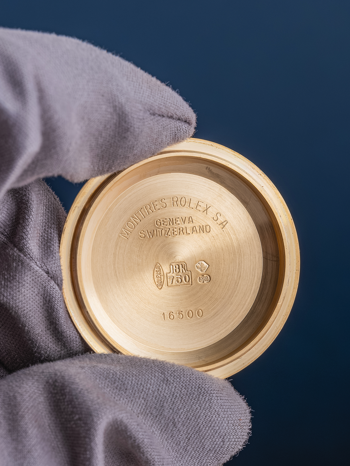 Gold watch case with 'Rolex' engraving held by a gloved hand against a blue background