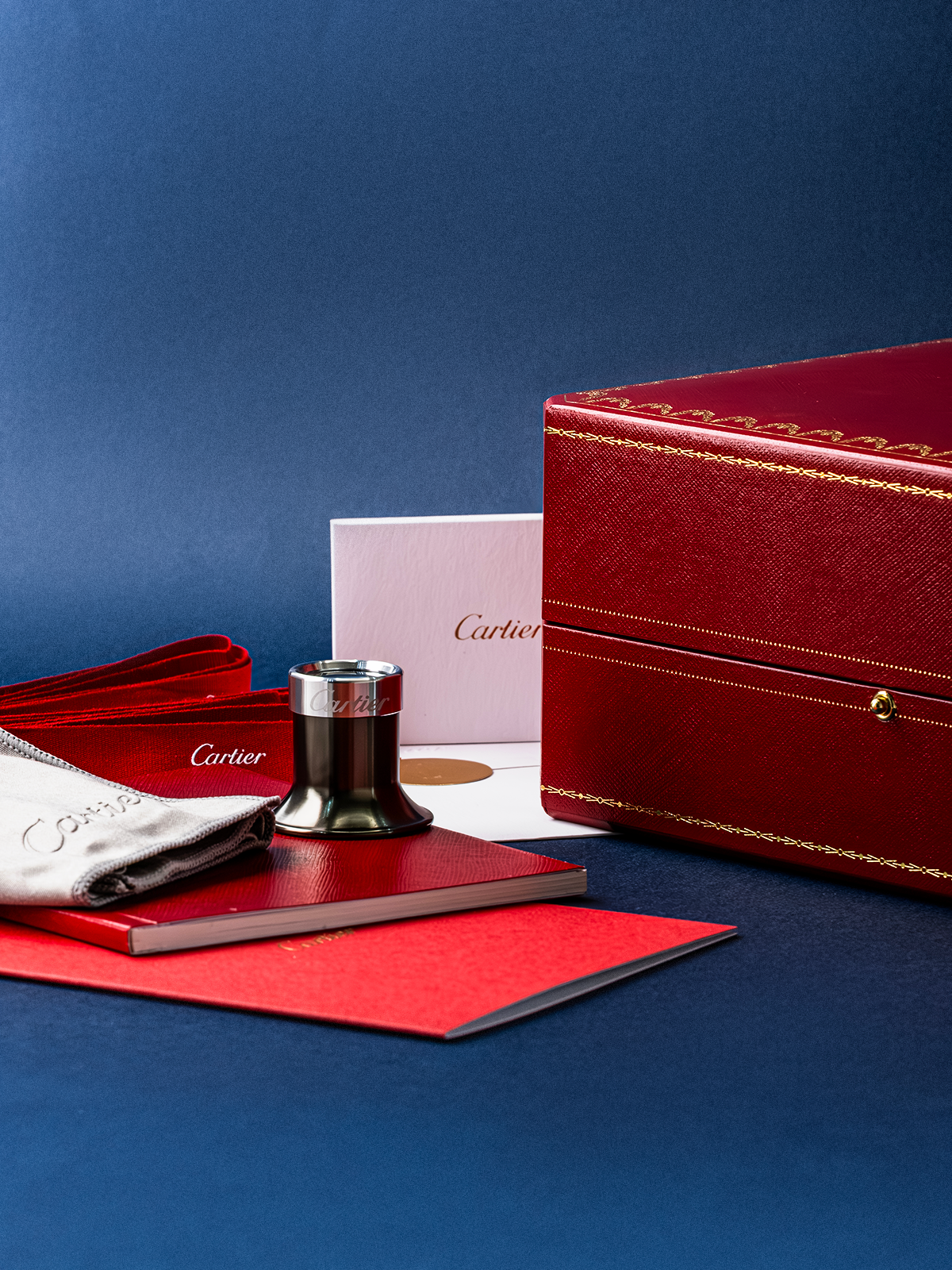 Cartier jewelry set with red box, silver cup, and pink card on a blue background