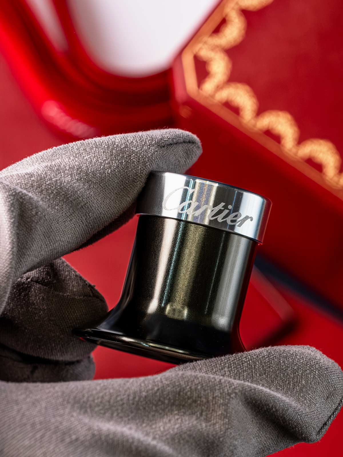 Black container with silver lid held by a gloved hand against a red background